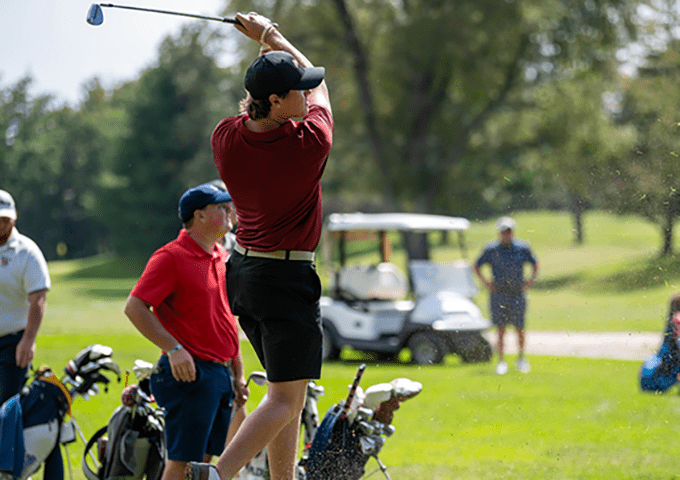 A golfer in a maroon shirt and black shorts is mid-swing with a driver or wood club, having just completed their swing.