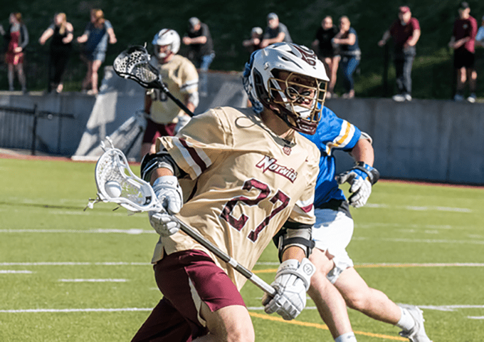 A player in gold and maroon #27 drives forward with the ball, closely guarded by an opponent in blue and yellow, with spectators blurred in the background.