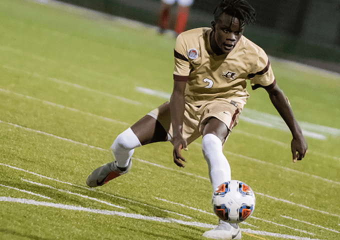 College soccer player in gold uniform dribbles the ball during a nighttime match on a green turf field, demonstrating precise ball control.