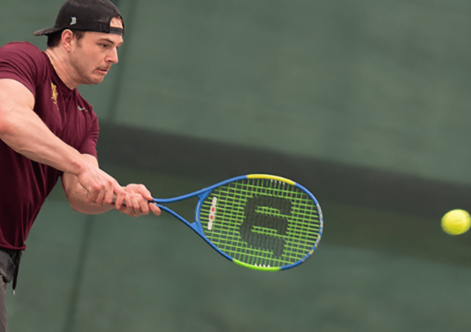 Male tennis player in maroon shirt and backwards cap hitting a forehand with blue and green racket, ball approaching racket against green court background.
