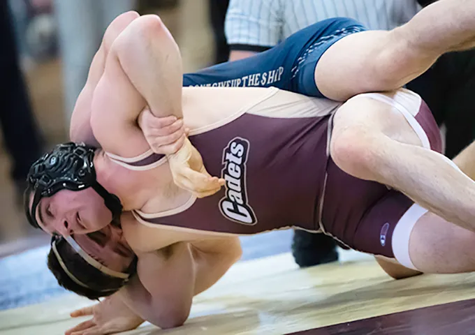 College wrestler in maroon singlet executes a pinning combination on the mat, demonstrating technical control over opponent during competition.