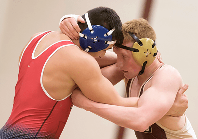 Two university wrestlers grapple in a match, with one competitor in a red singlet facing an opponent in a light-colored uniform with protective headgear.