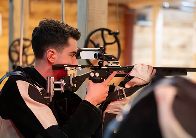 A person in a specialized shooting jacket (typically worn for stability in competitive shooting) aiming a competition rifle equipped with a target sight at an indoor shooting range.