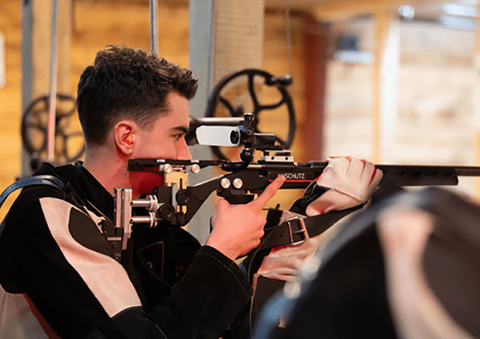 A person in a specialized shooting jacket (typically worn for stability in competitive shooting) aiming a competition rifle equipped with a target sight at an indoor shooting range.