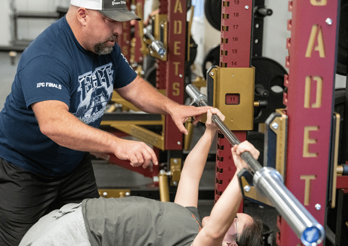 A coach spots a student athlete as they lift a bar while laying on a bench press in the weight room.