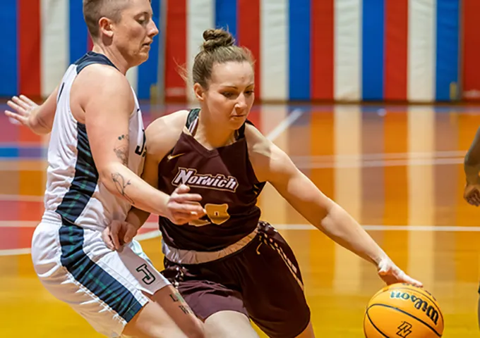 A Norwich basketball player in maroon and gold dribbles past a defender in white and blue on a polished court with a colorful backdrop.