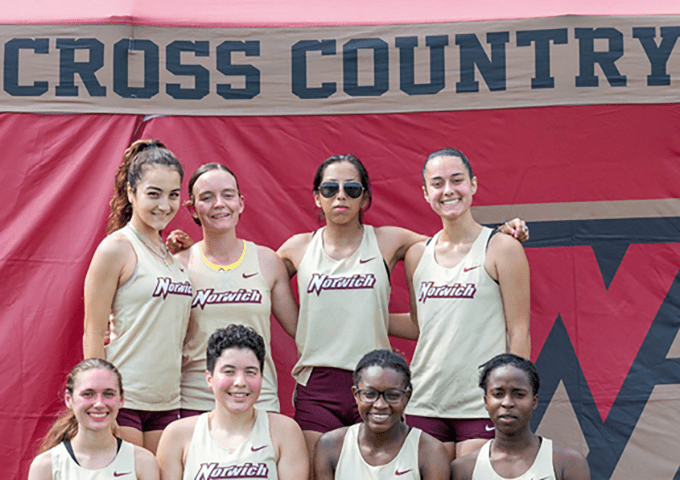 A group of six young women in the same jersey pose in front of a banner that reads "Cross Country".