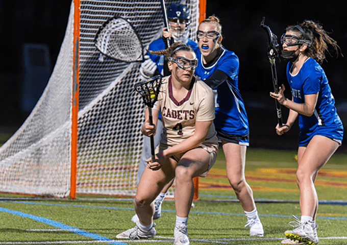 A lacrosse player in a cream and maroon uniform defending the goal against two opposing players in blue uniforms, with the net and goalie in the background.