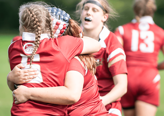 Rugby players in red jerseys celebrating together, one wearing an American flag bandana and braided hair, holding a trophy while embracing teammates on the field.