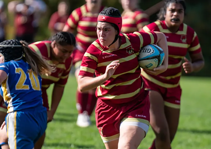 Rugby player in red and gold striped uniform carrying ball while running, wearing protective headgear, with teammates following and opponent in blue jersey nearby on grassy field.