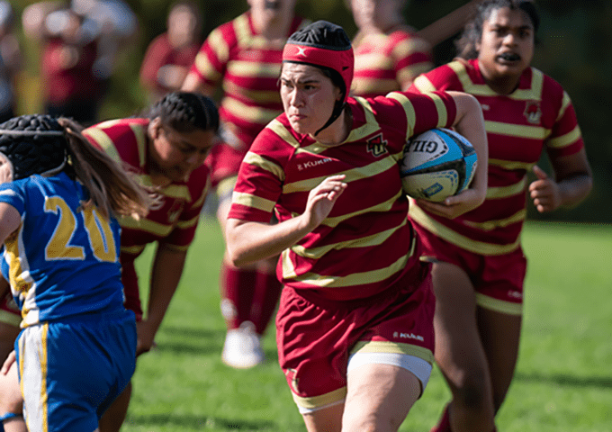 Rugby player in red and gold striped uniform carrying ball while running, wearing protective headgear, with teammates following and opponent in blue jersey nearby on grassy field.