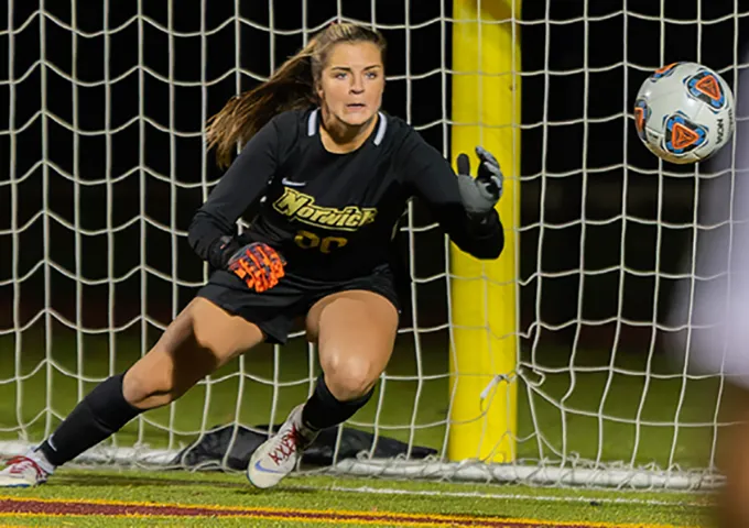 Focused goalkeeper in black uniform prepares to defend as a shot enters the net, demonstrating quick reaction in front of the goal posts.