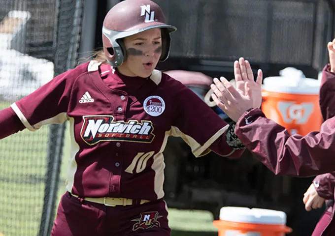 Norwich softball player in maroon uniform and batting helmet receiving high-five from coach near dugout, wearing number 10 jersey.