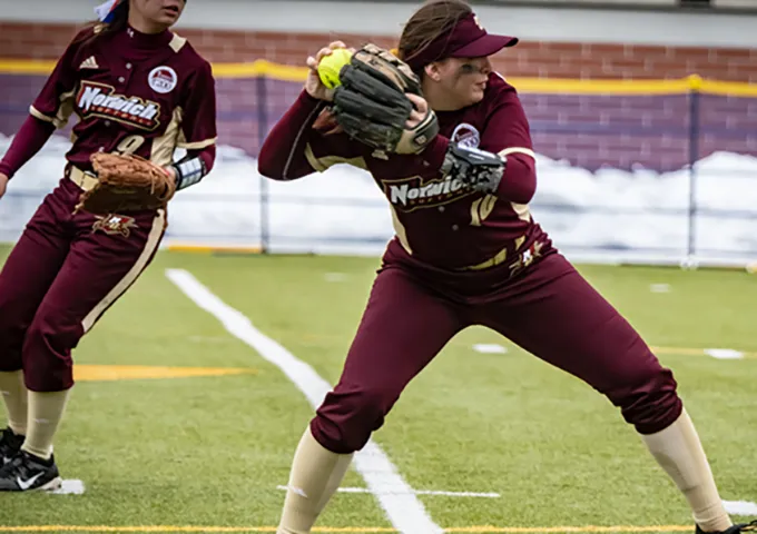 Norwich softball fielders in maroon uniforms with gloves ready; player in foreground in throwing stance while teammate stands behind on snowy field.
