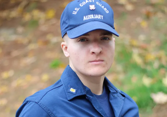 A close-up of a young adult with short, light hair wearing a blue U.S. Coast Guard Auxiliary cap and uniform shirt, looking neutrally at the viewer. The background is softly blurred autumn foliage.