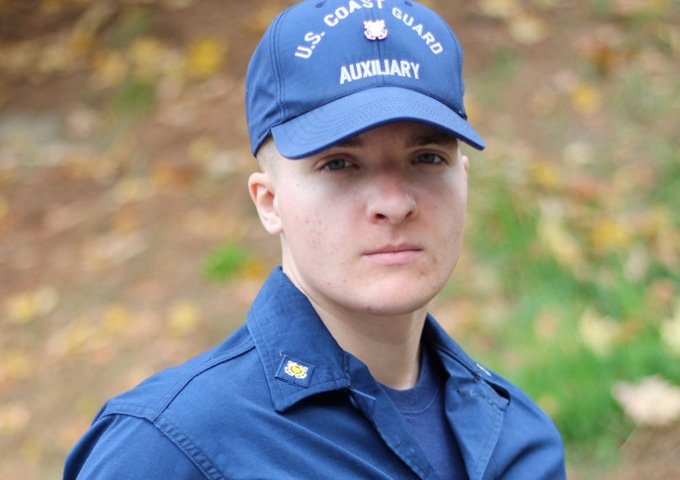 A close-up of a young adult with short, light hair wearing a blue U.S. Coast Guard Auxiliary cap and uniform shirt, looking neutrally at the viewer. The background is softly blurred autumn foliage.