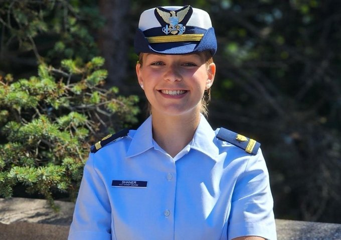 U.S. Coast Guard officer in white uniform shirt and blue cap smiling with arms crossed, standing in front of brick wall with evergreen trees in background.