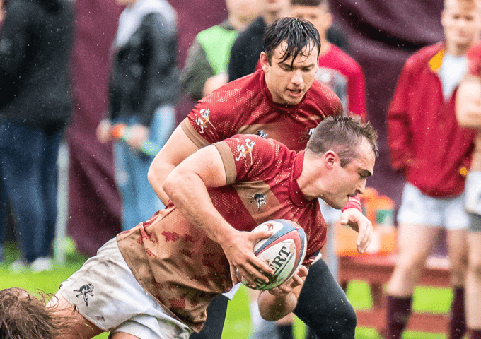 Rugby player in red drives forward with the ball in rainy conditions as a teammate supports from behind during a tense play.