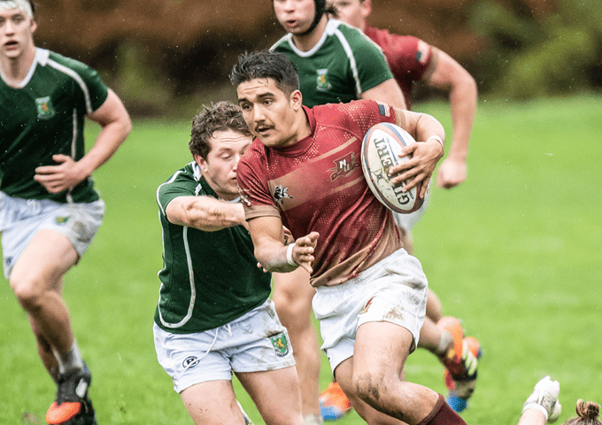 Rugby player in red breaks a tackle from a defender in green during a wet, grassy field match.