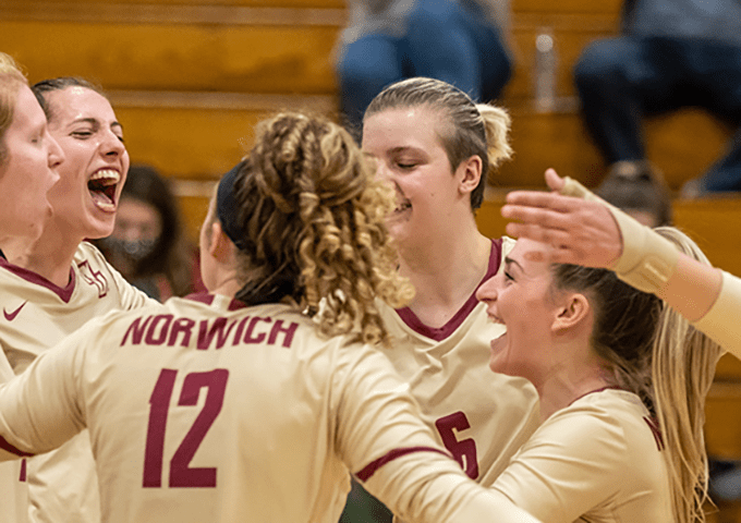 Volleyball team members in tan jerseys celebrating together on court, with player number 12 visible from behind.