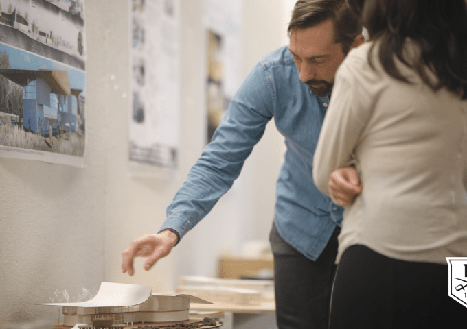 Person in a blue shirt gestures to an architectural model on a table while another individual listens. Design posters are displayed on the wall; Norwich emblem visible.