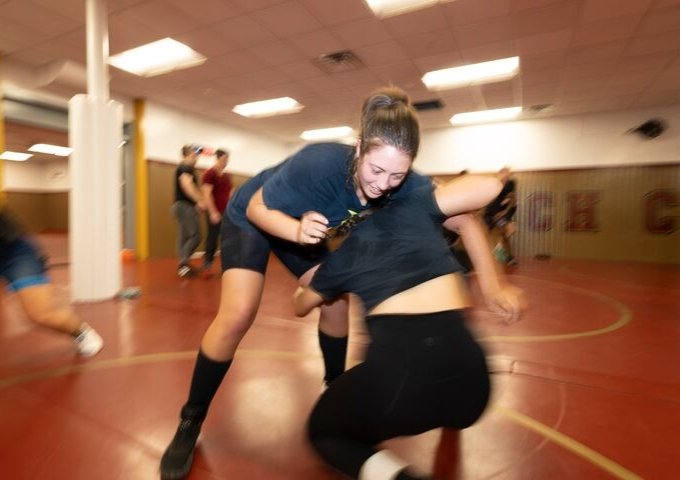 Two people on a red mat wrestle, with one grabbing the other's waist; blurred figures stand in the background.