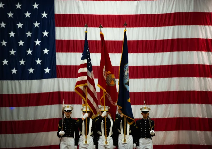 Military honor guard stands in front of a large American flag, holding three flags.
