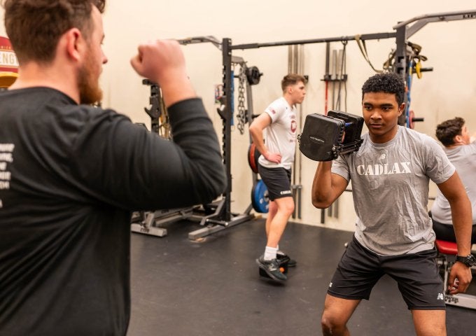 Athlete in a CADLAX shirt performs a weighted squat in a gym while a trainer gestures and others lift in the background.