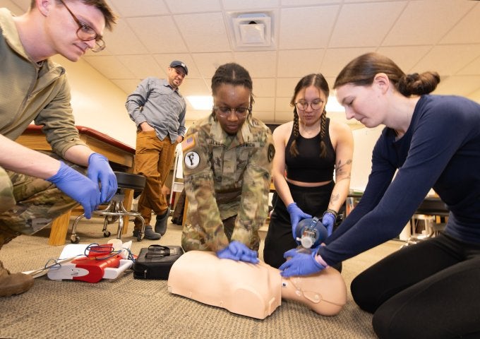 Group of students and a uniformed cadet practice CPR on a training mannequin while another uses a defibrillator nearby.