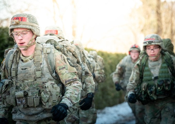 Service members in camouflage uniforms carry heavy packs while marching in a line during outdoor training.