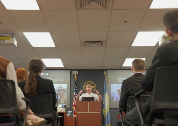 Person speaks at a podium to seated audience in a room with flags and presentation slides on screens.