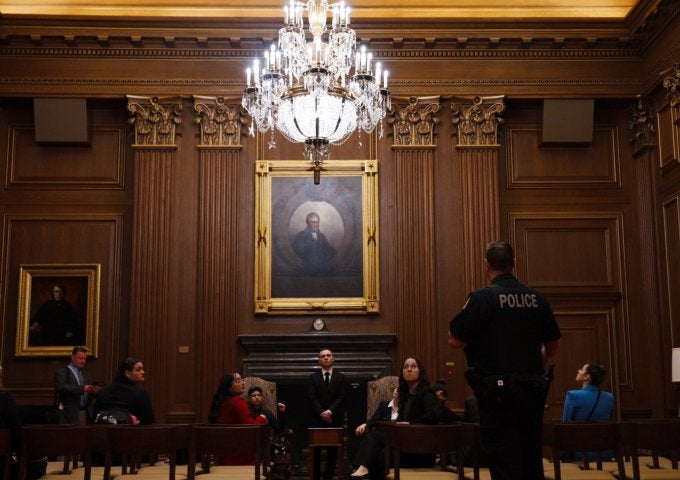 Police officer stands before seated people in a wood-paneled courtroom with a chandelier and framed portraits.