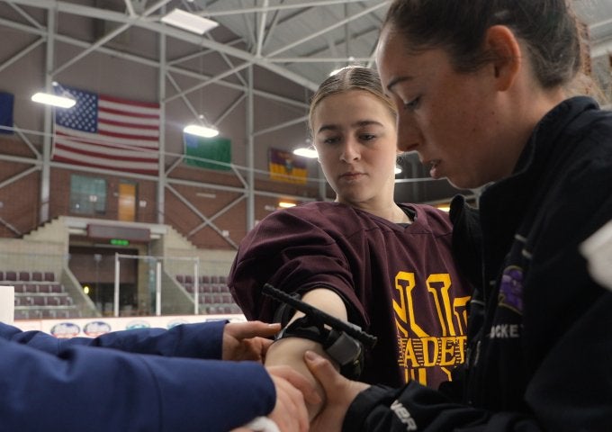 Hockey player in a Norwich Cadets jersey has an arm brace adjusted by staff beside an indoor ice rink.