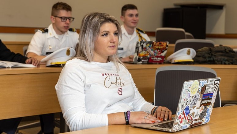 Civilian student on laptop sitting in classroom with two cadets sitting behind.