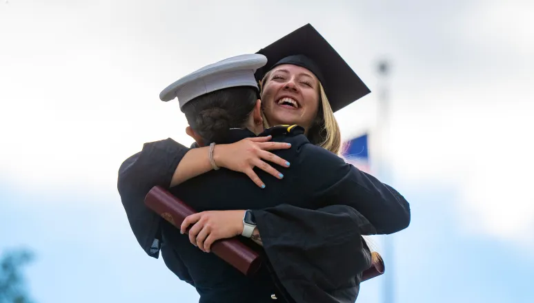 A corps and civilian student in graduation garb embracing with diploma in hand.