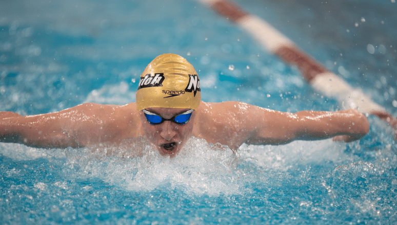 Norwich swimmer wearing a gold cap and blue goggles powers through the water in a butterfly stroke, creating a splash.