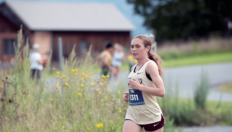 Runner in Norwich jersey, number 1311, racing past yellow wildflowers and a rural building.