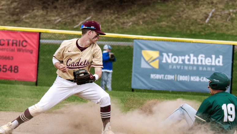 Baseball player in a gold 'Cadets' jersey prepares to tag out a sliding opponent wearing a green uniform with number 19, as dust rises around them near second base.