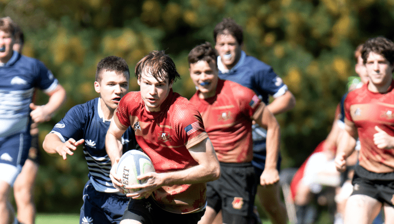 Rugby player in red evades defenders in blue, sprinting with the ball during a sunny outdoor match.