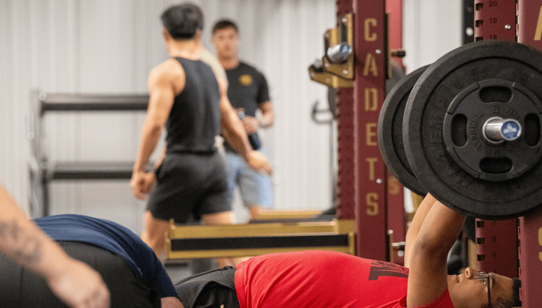 Weightlifting session in a gym with barbell rack and weight plates; people training in a cadets fitness facility.