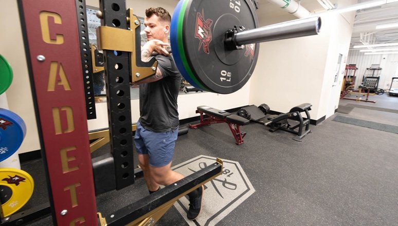 Person in athletic wear performing a front squat with a loaded barbell in the Norwich University 84 Fitness Center weight room.