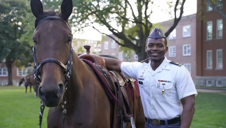 Cadet in uniform stands beside a saddled horse on campus.