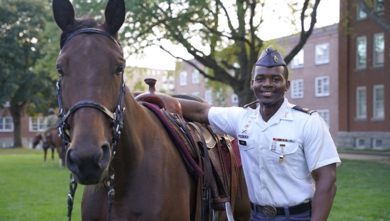 Cadet in uniform stands beside a saddled horse on campus.