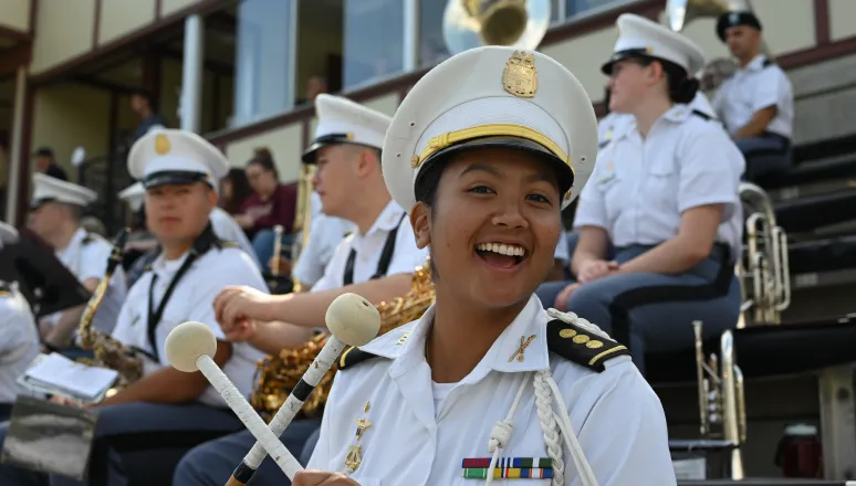 A smiling band member in white uniform and hat holds mallets, with other musicians in white uniforms seated behind.