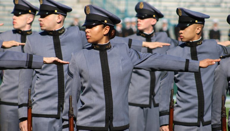 Line of cadets in gray dress uniforms and caps performing a synchronized arm movement during outdoor drill practice.