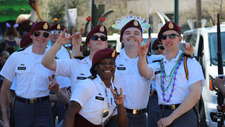 Group of cadets in white short-sleeve uniforms laughing and posing together, some wearing colorful beads and costume accessories.
