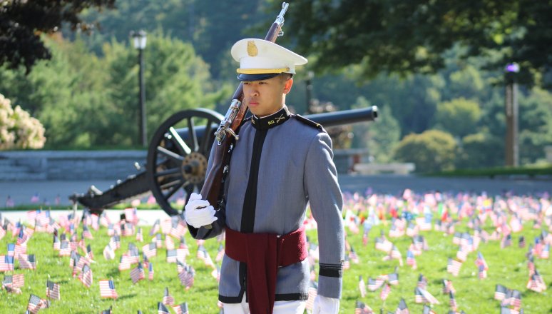 Cadet in gray dress uniform and white hat stands at attention with a rifle over one shoulder on a lawn filled with small American flags and a cannon in the background.