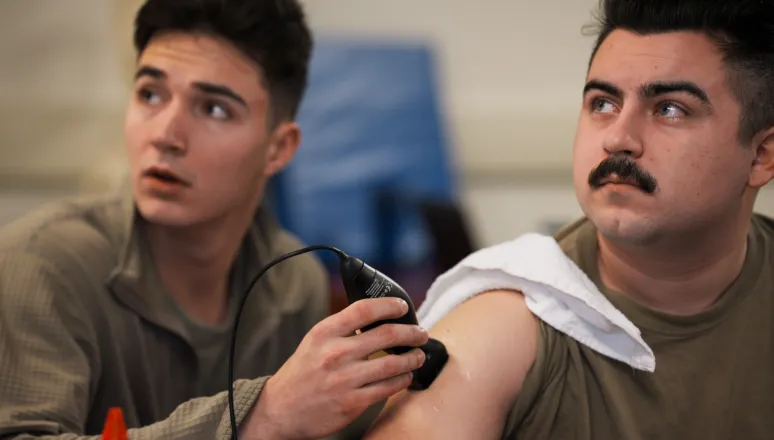 Student uses a handheld ultrasound device on another student's upper arm during a classroom training session.