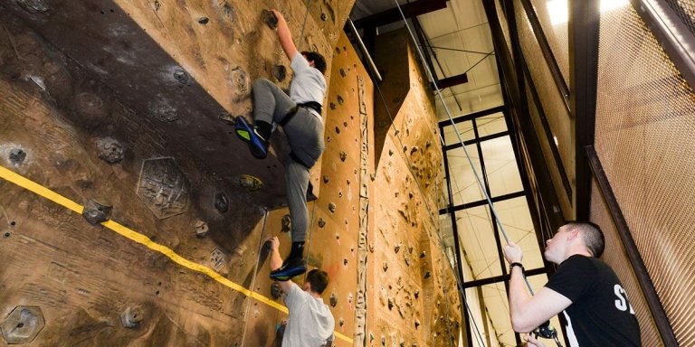 Person climbing an indoor rock wall with a harness, while another person on the ground manages the safety rope as the belayer.