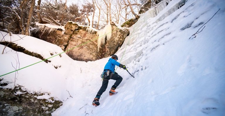 Person ice climbing a snowy, steep slope using crampons and ice axes, secured with a rope in a forested winter landscape.